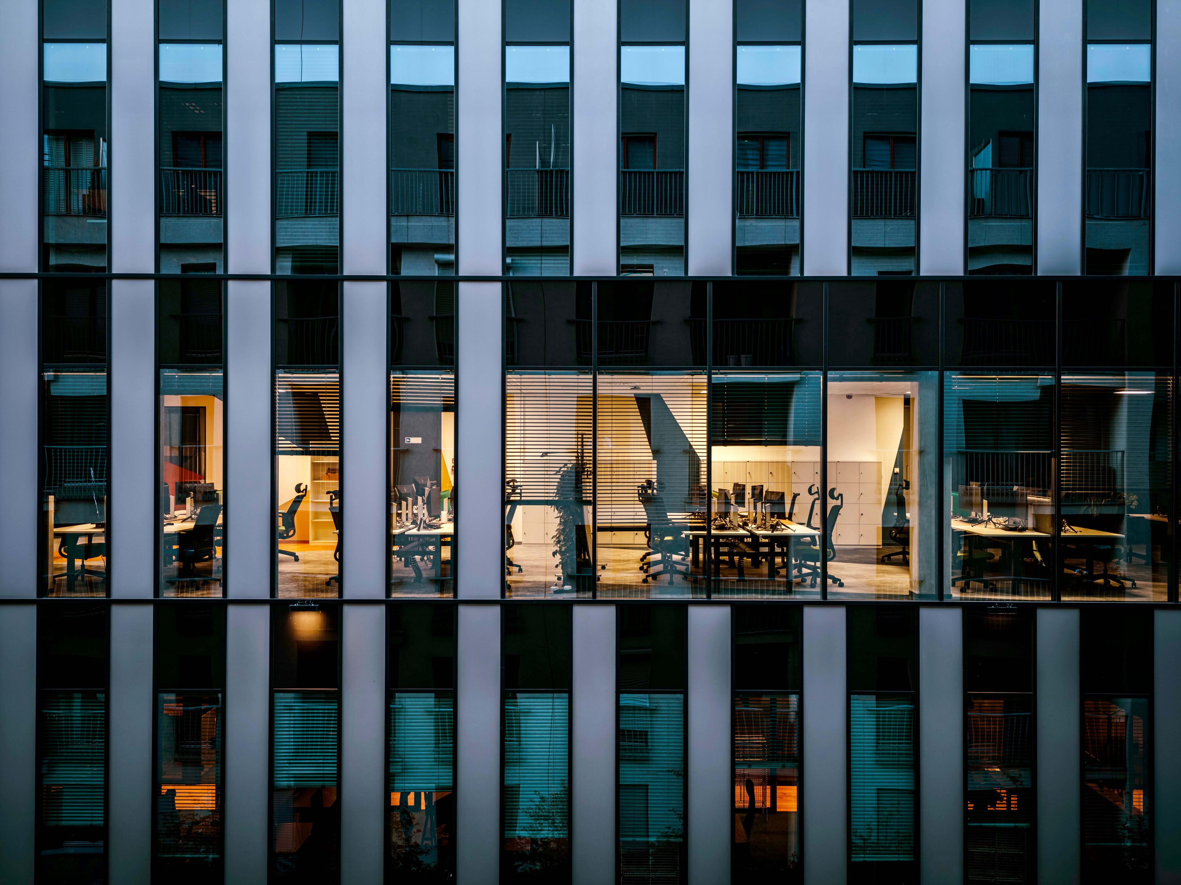 Lit offices seen through the glass facade of an office building at dusk